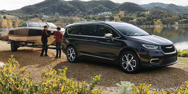 Black Chrysler Pacifica minivan parked by a lakeside area with people preparing a boat on a trailer.