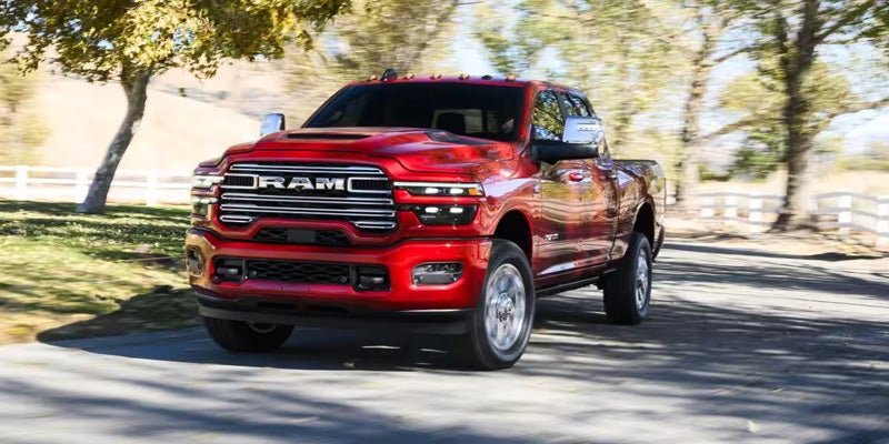 2026 Ram 2500 red heavy-duty pickup truck driving on a tree-lined road, showcasing its bold front grille and rugged design.